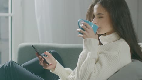 Beautiful-Young-Woman-Using-Smartphone,-Drinks-Tea-while-Sitting-on-the-Chair.-Sensual-Girl-Wearing-Sweater-Surfs-Internet,-Posts-On-Social-Media,-Shares-Pictures-while-Relaxing-in-Cozy-Apartment