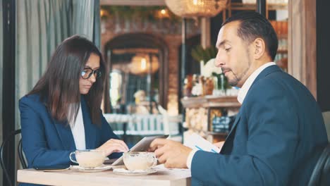 Two-business-people-have-business-lunch.-The-man-and-woman-in-a-restaurant-or-a-coffee-shop-are-drinking-coffee,-and-woman-is-working-on-tablet.-Man-is-looking-through-documents.-Copy-space.