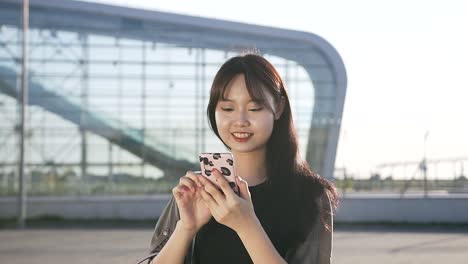 Beautiful-smiling-young-asian-woman-walking-near-airport-building-and-using-her-mobile