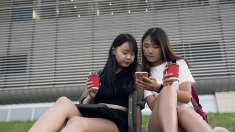 Atractivas-chicas-asiáticas-sonrientes-felices-sosteniendo-en-las-manos-café-y-mirando-el-teléfono,-sentado-en-la-hierba-cerca-de-un-gran-edificio-urbano