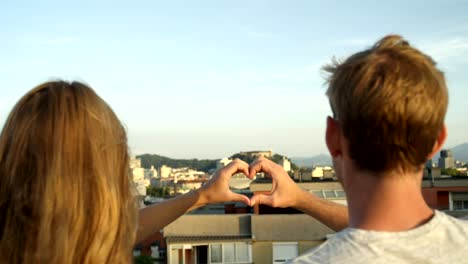 CLOSE-UP:-Pareja-haciendo-el-símbolo-en-forma-de-corazón-y-besos-en-azotea-sobre-ciudad