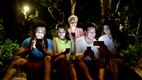 Personas-grupo-usando-teléfono-inteligente-celular-sentado-en-el-banco-en-el-parque-de-la-noche,-jóvenes-amigos-hablando-de-redes-en-línea