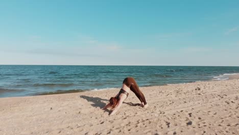 Drone-bewegt-sich-hinter-Frau-in-Sport-Wear-hält-Yoga-Asana-Position-am-Meer-oder-Ozean-Sandstrand.-Windige-sonniges-Wetter.-Luftaufnahme-des-friedlichen-Gesundheit-Mädchen-Praxis-durchführen