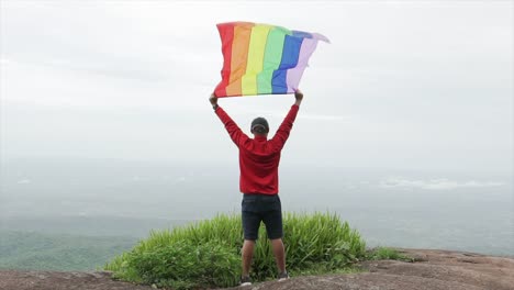 hombre-levantar-bandera-LGBTI-de-color-arco-iris-ondeando-en-duro-viento-en-el-mirador-de-alta-montaña