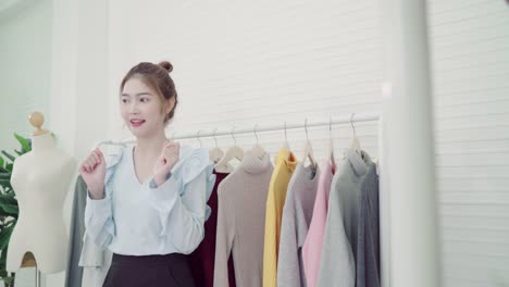 Asian-fashion-female-blogger-online-influencer-holding-shopping-bags-and-lots-of-clothes-on-clothes-rack-for-recording-new-fashion-video-broadcast-live-video-to-social-network-by-internet-at-home.