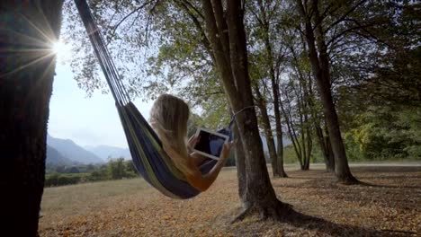 Female-in-hammock-reading-on-digital-tablet-using-mobile-apps-on-wireless-technology.-Young-woman-enjoying-nature-in-late-Summer-swinging-between-trees