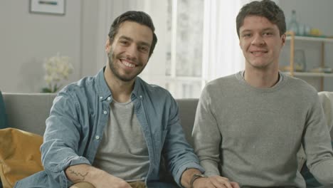 Pareja-Gay-masculino-atractivo-lindo-sentarse-juntos-en-un-sofá-en-casa.-Novio-pone-su-mano-en-pareja.-Ellos-son-feliz-y-sonriente.-Son-casualmente-vestida-y-su-habitación-tiene-un-Interior-moderno.