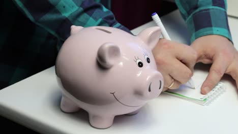 On-the-table-there-is-a-piggy-bank-in-the-form-of-a-pink-pig.-A-man-puts-a-coin-in-it,-makes-notes-in-a-notebook.