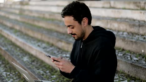 Young-Arabic-man-texting-on-phone,-holding-laptop-on-knees