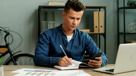 Joven-hombre-de-negocios-usando-teléfono-inteligente-fot-señalando-algo-en-el-libro-de-notas-mientras-trabajaba-en-la-oficina.