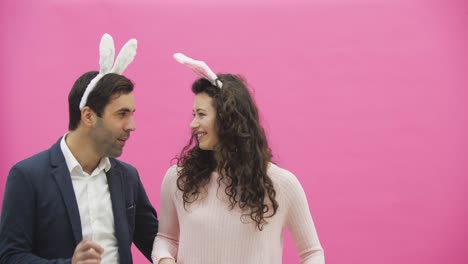 Young-beautiful-handsome-happy-happy-couple-standing-on-a-pink-background.-At-the-same-time-fools-showing-various-movements,-a-man-sings-his-wife-gently-looking-at-her.