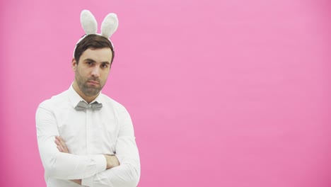 Young-handsome-man-standing-on-a-pink-background.-Dressed-in-a-white-shirt-with-rabbit-ears-on-his-head.-Putting-a-hand-on-the-hand,-serious,-looking-into-the-camera.