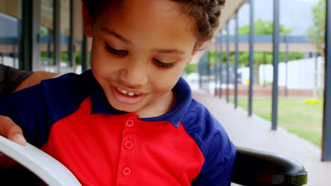 Front-view-of-disabled-African-American-schoolboy-reading-a-book-on-wheelchair-in-school-corridor-4k