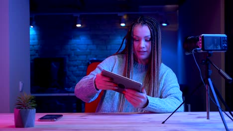 Closeup-shoot-of-young-attractive-female-blogger-with-dreadlocks-texting-on-the-tablet-streaming-live-with-the-neon-background-indoors