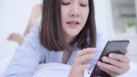 Junge-asiatische-Frau-mit-Smartphone,-während-auf-dem-Bett-nach-dem-Aufwachen-am-Morgen,-Schöne-attraktive-japanische-Mädchen-lächelnd-entspannen-im-Schlafzimmer-zu-Hause.-Spaß-im-Lifestyle-Frauen-zu-Hause-Konzept.