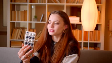 Young-teenage-blogger-talking-in-videochat-on-cellphone-sitting-on-sofa-on-bookshelves-background-at-home.