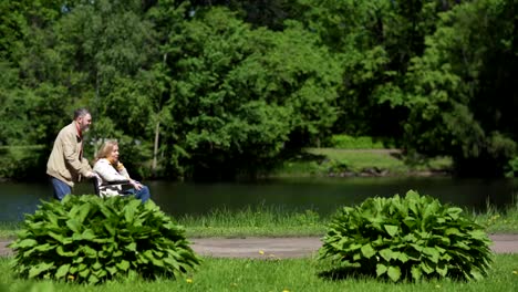 Lockdown-Seitenansicht-von-Senior-Mann-schiebt-seine-behinderte-Frau-im-Rollstuhl-auf-dem-Weg-in-der-Nähe-von-Teich-in-grünen-Park.-Reife-Ehepartner-im-Gespräch