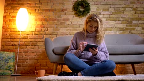 Closeup-top-shoot-of-adult-caucasian-blonde-female-using-the-tablet-while-sitting-on-the-floor-indoors-in-a-cozy-apartment