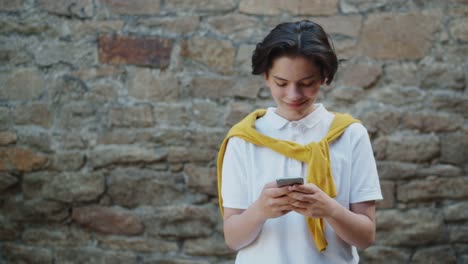 Smiling-teenage-kid-touching-smartphone-screen-standing-alone-in-the-street