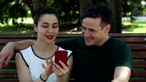 A-young-beautiful-girl-and-her-handsome-young-boyfriend-watch-the-news-in-the-social-network-on-their-smartphone-while-sitting-on-a-park-bench.