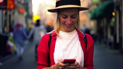 Atractiva-mujer-caucásica-en-camisa-casual-roja-escribiendo-mensaje-de-texto-en-el-teléfono-móvil-mientras-está-de-pie-al-aire-libre