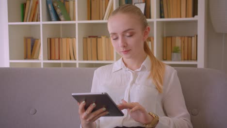 Closeup-portrait-of-young-pretty-caucasian-blonde-female-student-using-the-tablet-looking-at-camera-smiling-sitting-on-the-couch-indoors-in-the-apartment