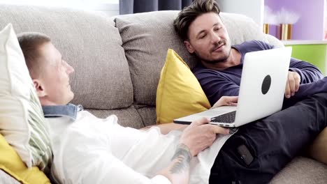 Gay-couple-relaxing-on-couch-using-laptop-computer.-Talking.