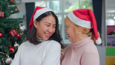 Pareja-lesbiana-asiática-celebran-festival-de-Navidad.-LGBTQ-adolescente-mujer-usan-sombrero-de-Navidad-relajarse-feliz-sonriendo-mirando-a-la-cámara-disfrutar-de-las-vacaciones-de-invierno-de-Navidad-juntos-en-la-sala-de-estar-en-casa.