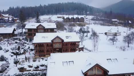 Aire-de-cabañas-de-madera-en-pueblo-de-montaña-rodeado-de-bosque-de-coníferas.-Vista-de-drone-sin-chalets-cubiertos-de-nieve-en-la-estación-de-esquí.-Frío-día-de-invierno-helado-y-nevadas-en-las-montañas