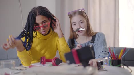 Chica-afroamericana-con-anteojos-rojos-y-su-amiga-caucásica-con-bigotes-falsos-sentados-en-la-mesa-y-mirando-la-pantalla-del-teléfono-inteligente.-Adolescentes-tomando-selfie-foto.-Diversión,-felicidad,-estilo-de-vida.