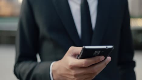 Handsome-asian-businessman-in-a-black-suit-using-his-smartphone-browsing-internet,-scrolling-social-media-while-standing-outside-on-the-street-near-a-big-office-building-urban-having-sunlight-sunset.