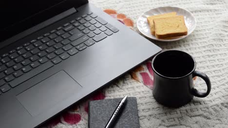 Café-de-desayuno-a-la-luz-del-sol-de-la-mañana-con-pluma-de-color-negro-computadora-portátil-y-cuaderno-organizador-personal,-platillo-de-taza-de-cerámica-y-galleta-en-el-fondo-de-escritorio-del-lugar-de-oficina-superior.-Imagen-de-estilo-de-vida.