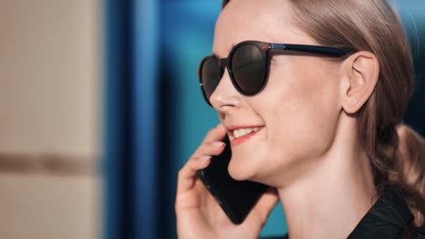 Cara-de-primer-plano-de-la-mujer-jefa-sonriente-en-gafas-de-sol-hablando-smartphone.-Grabado-con-cámara-RED-en-4K