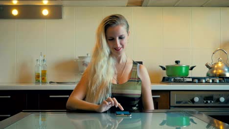 Dolly-the-young-girl-sits-at-the-table-and-read-on-the-smartphone-in-the-kitchen-at-home.