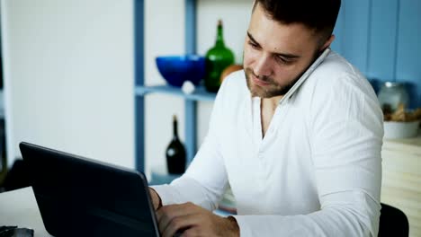 Handsome-young-man-talking-phone-and-using-laptop-computer-sitting-in-the-kitchen-after-breakfast-in-the-morning