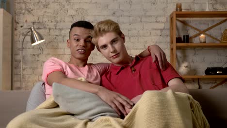 Two-gay-guys-are-sitting-on-the-couch-and-watching-TV,-a-Caucasian-guy-wants-to-switch-the-channel-but-an-African-guy-stops-him.-LGBT-lovers,-a-multinational-couple,-a-happy-gay-family,-a-home-cosiness-concept.-Look-at-the-camera-60-fps