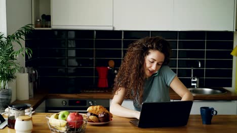 Joven-rizado-bonita-a-mujer-caucásica-positiva-trabajando-en-equipo-portátil-y-hablando-por-teléfono-en-cocina,-es-impresión-y-hablar-al-mismo-tiempo