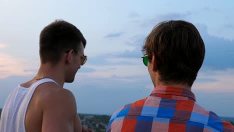 Dolly-shot-of-young-male-couple-in-sunglasses-standing-on-the-edge-of-rooftop-and-talking.-Handsome-gay-boys-rest-on-roof-of-high-rise-building-and-enjoying-beautiful-cityscape.-Close-up-Slow-motion