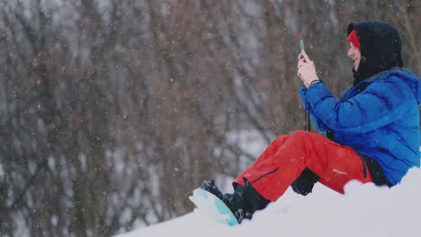 Snowboarder-masculino-sentado-en-la-nieve-tomando-fotos-en-el-teléfono-hermoso-paisaje-del-complejo-para-las-redes-sociales