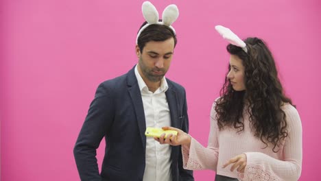 Young-lovers-couple-on-the-pink-background.-With-hackneyed-ears-on-the-head.-During-this,-the-photos-of-Sephi-on-the-phone-are-very-sincere-smiles.-Funny-people.