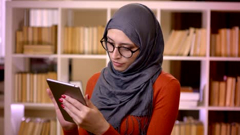 Closeup-shoot-of-young-attractive-muslim-female-student-in-hijab-typing-on-the-tablet-standing-indoors-in-the-library