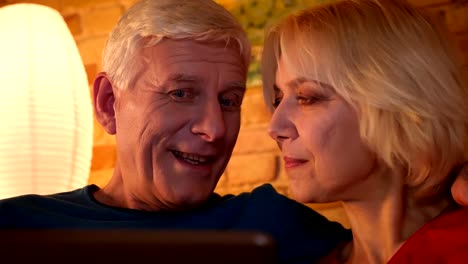 Closeup-shoot-of-aged-happy-couple-browsing-the-network-on-the-tablet-sitting-on-the-sofa-resting-together-indoors-in-a-cozy-flat