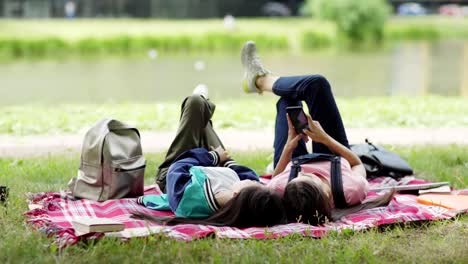 Toma-de-seguimiento-de-dos-amigos-relajados-de-la-universidad-descansando-en-el-césped-cerca-del-estanque-después-de-las-clases.-Las-alumnas-tumbadas-en-la-manta,-navegando-por-Internet-en-el-teléfono-inteligente-y-hablando