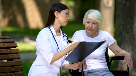 Médico-femenino-que-muestra-resultado-de-rayos-X-espinal-a-la-anciana-discapacitada,-día-en-el-parque