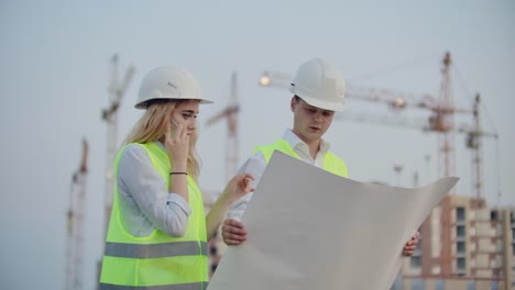 Woman-talking-on-the-phone-and-asks-the-Builder-what-is-on-the-drawings-standing-on-the-background-of-buildings-under-construction