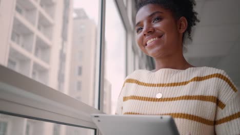 Porträt-einer-glücklichen-jungen-afrikanischen-Geschäftsfrau,-die-mit-einem-digitalen-Tablet-in-der-Nähe-des-Bürofensters-steht