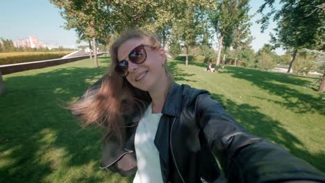 Female-student-in-the-park.-Charming-girl-in-leather-jacket-walks-around-autumn-park-and-films-herself-on-the-camera.-Makes-a-selfie-on-a-smartphone-for-social-networks.-slow-motion.