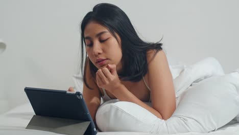 Junge-asiatische-Frau-mit-Tablet-Checking-Social-Media-Gefühl-glücklich-lächeln,-während-auf-dem-Bett-liegen,-nachdem-sie-morgens-im-Haus-aufwachen,-Attraktive-indische-weibliche-lächelnd-entspannen-im-Schlafzimmer-zu-Hause-Konzept.