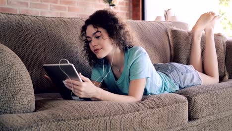 Hispana-de-pelo-rizado-joven-mujer-navegando-por-la-red-usando-tableta-digital-y-escuchando-música-mientras-se-relaja-en-un-sofá