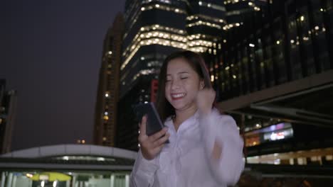 Emocionada-mujer-asiática-de-negocios-celebrando-buenas-noticias-revisando-el-teléfono-inteligente-en-el-centro-de-la-ciudad-moderna-en-la-noche-de-Bangkok-Tailandia.-Comunicación-Concept-Technology,-éxito-por-teléfono-móvil